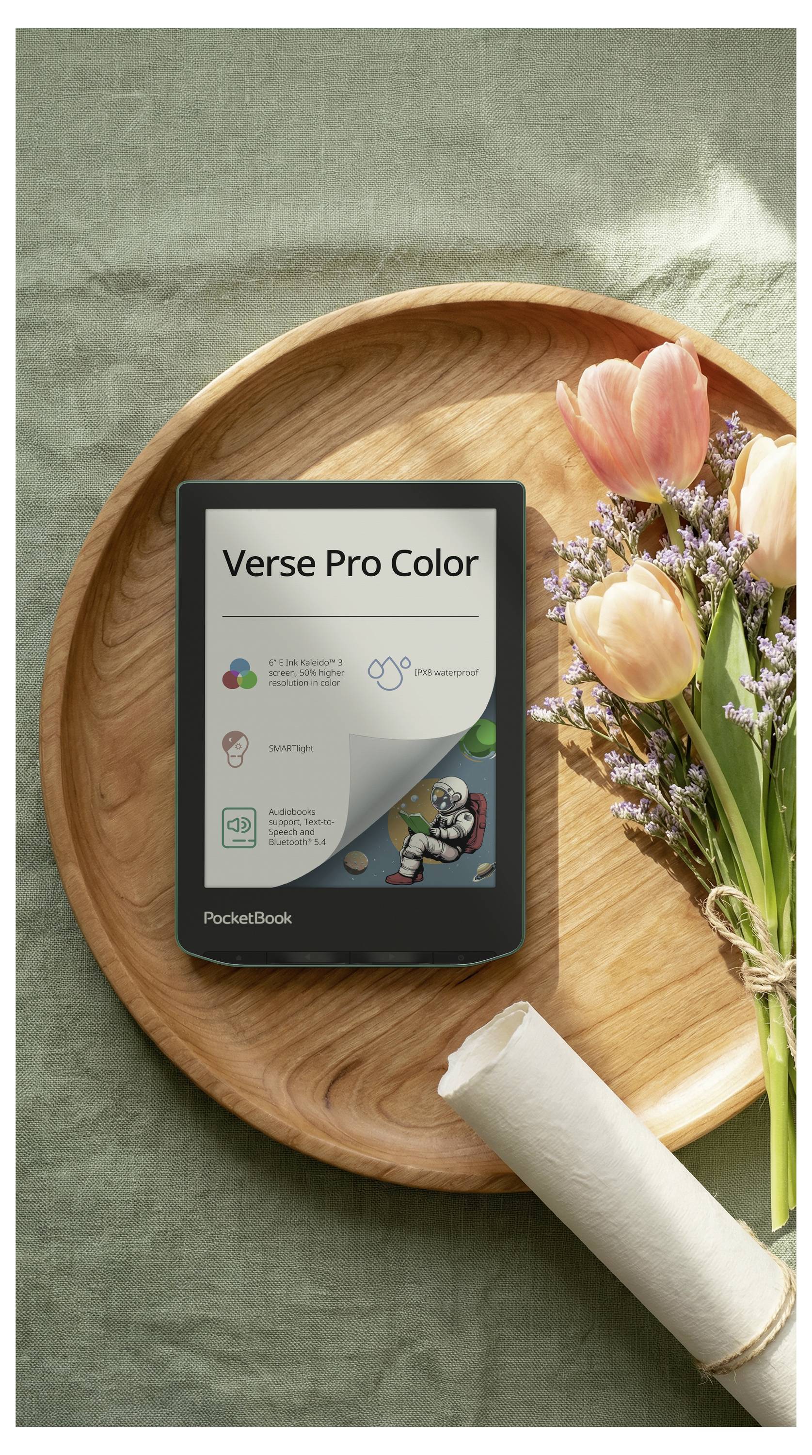 PocketBook Verse Pro Color e-čitač na drvenom pladnju uz cvijeće i umotanu salvetu na zelenoj tkanini u pozadini.
