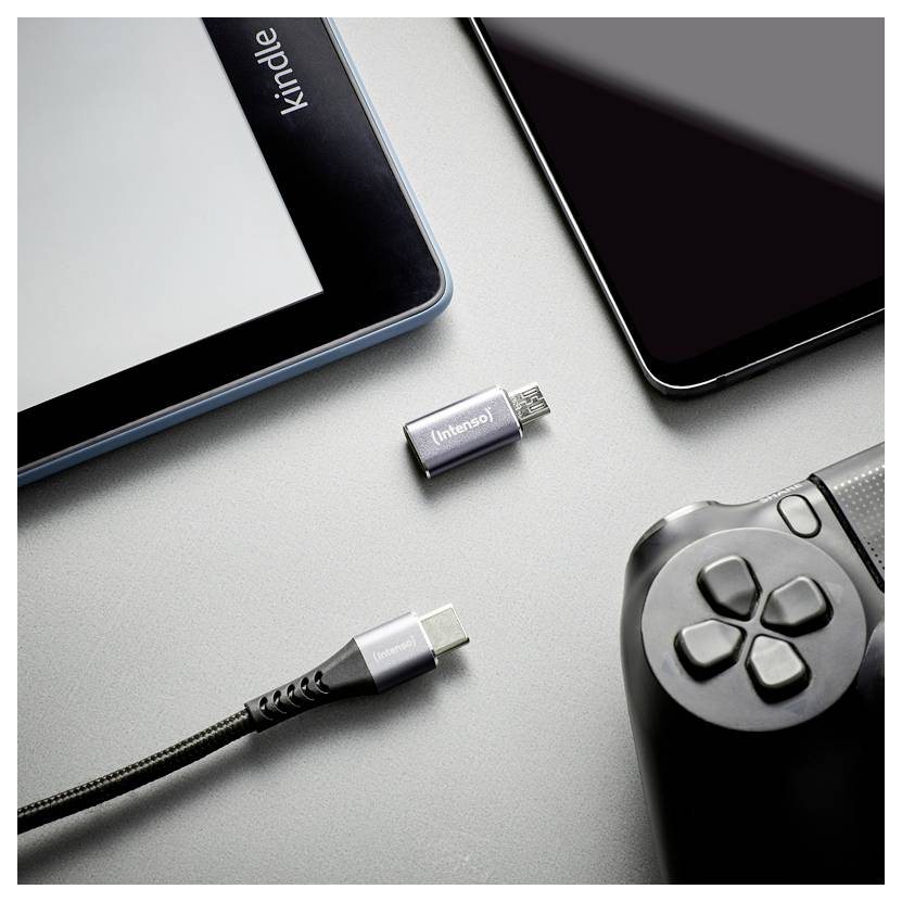 USB-C adapter i kabel pored tableta, pametnog telefona i kontrolera za igre na sivoj površini, što sugerira punjenje ili povezanost uređaja.