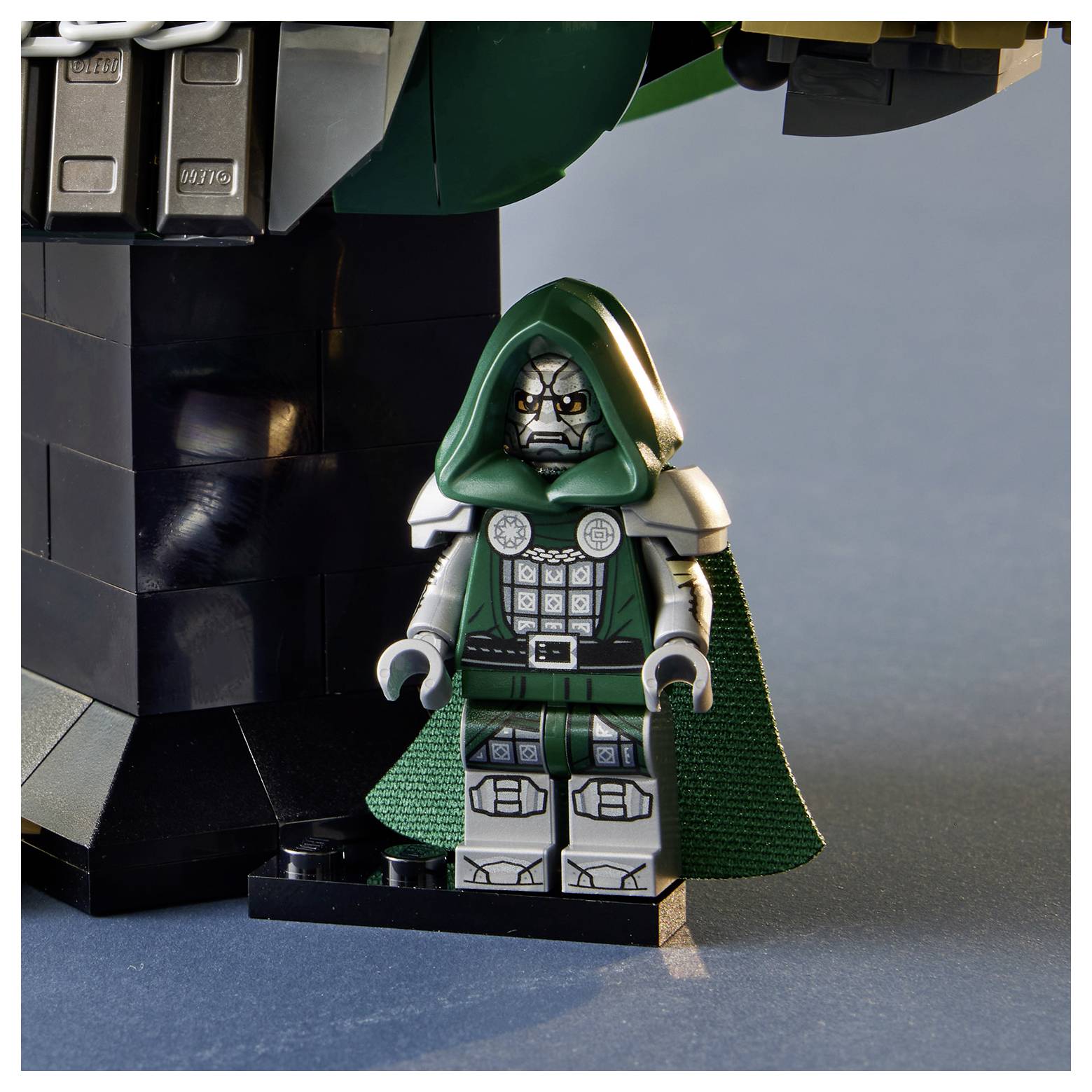 Minijatura LEGO Doktora Dooma u zelenoj kapuljači stoji pored većeg crnog LEGO objekta na plavoj površini.
