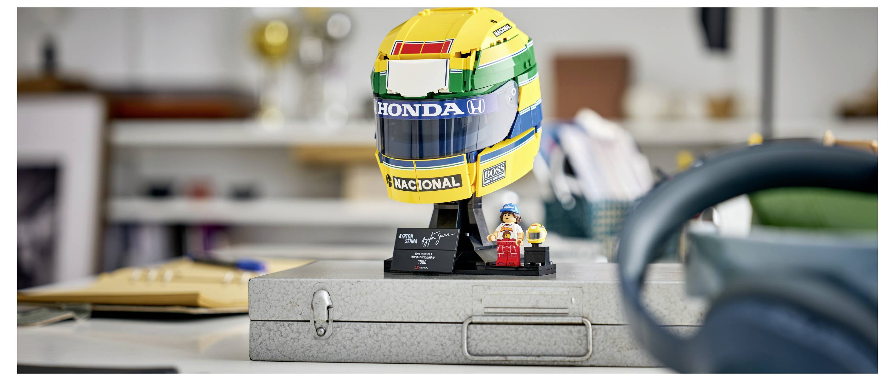Minijaturna LEGO trkaća kaciga s natpisima "HONDA" i "NACIONAL" nalazi se na sivoj kutiji s alatima, pored male minifigurice u kancelarijskom okruženju.