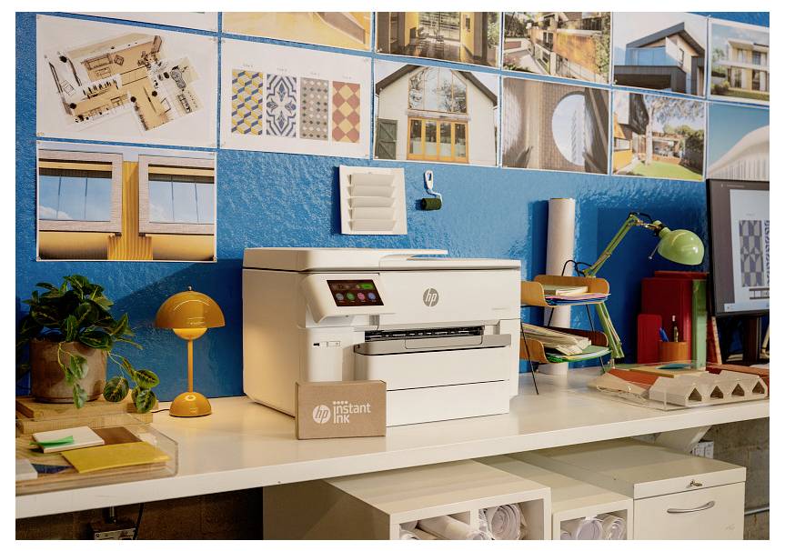 HP Instant Ink printer na stolu u svjetloj radnoj prostoriji, s biljkom, lampom, priborem i arhitektonskim fotografijama prikupljenim na plavoj zidu iza njega.