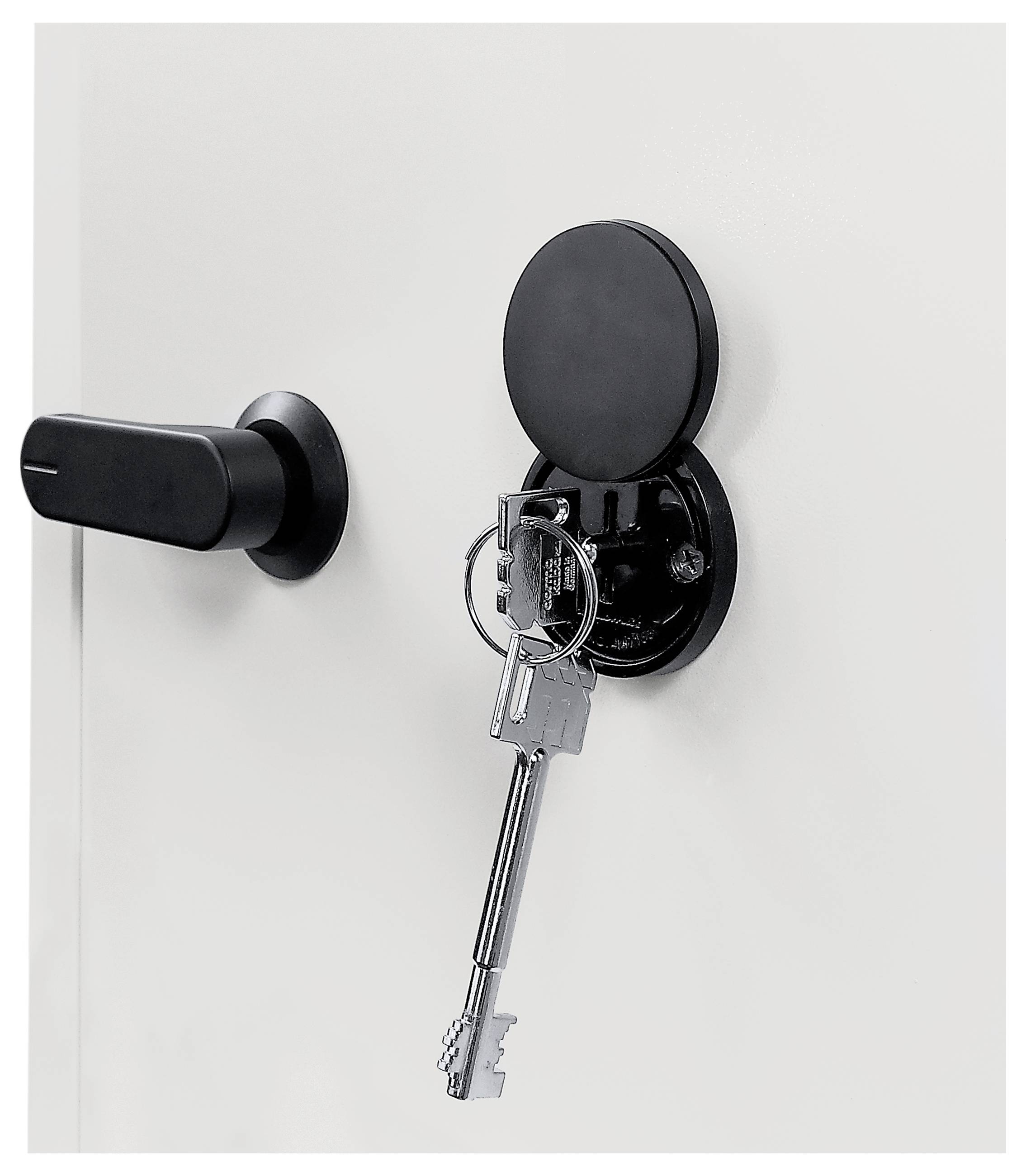 Crni ormarić za ključeve montiran na bijelim vratima, otvoren s dva ključa obješena na bravu pored crne ručke.