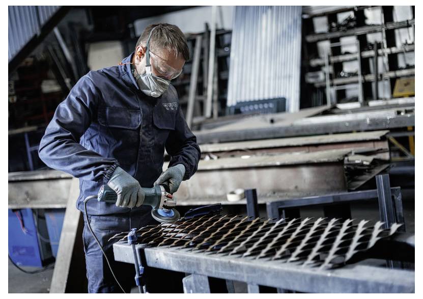 Radnik u zaštitnim naočalama i rukavicama koristi kutnu brusilicu na metalnoj mrežastoj tablici u industrijskoj radionici.