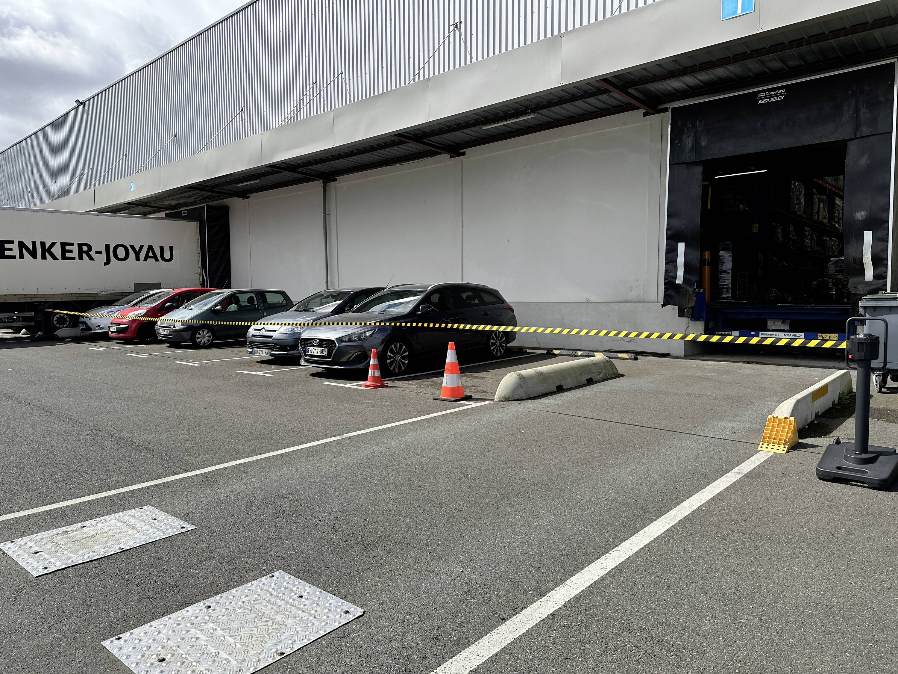 Automobili parkirani pored rampe za utovar u skladištu, blokirani crno-žutom opreznom trakom i prometnim čunjevima blizu otvorenih vrata balkona.