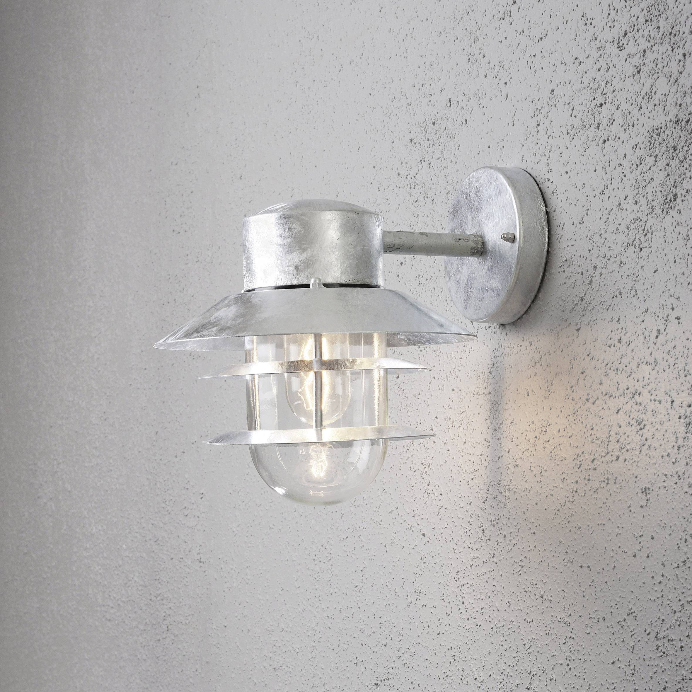 Zidna svjetiljka od metala s staklenim poklopcem, pričvršćena na sivom zidu. Lampa je uključena i odašilja toplo svjetlo.