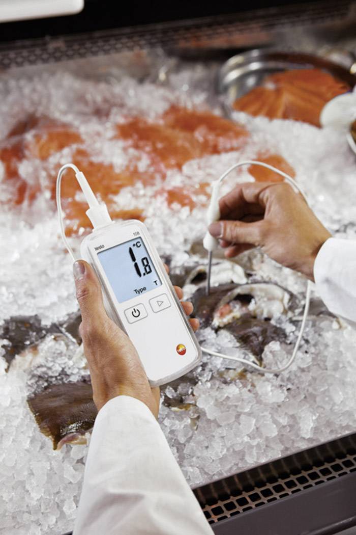 Jedna osoba mjeri temperaturu ribe koja leži na ledu u izlogu digitalnim termometrom. Display pokazuje 1,8 °C.