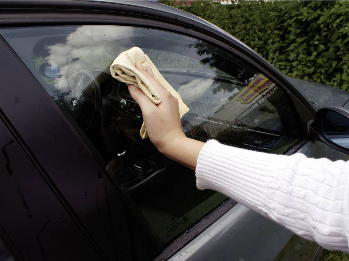 Jedna osoba čisti prozor automobila izvana bež krpom. Auto se nalazi vani, a oblačno nebo reflektira se u staklu.