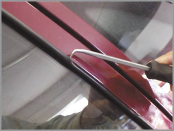 Jedna osoba koristi metalni alat kako bi povukla brtvilo prozora automobila. Automobil je crven.