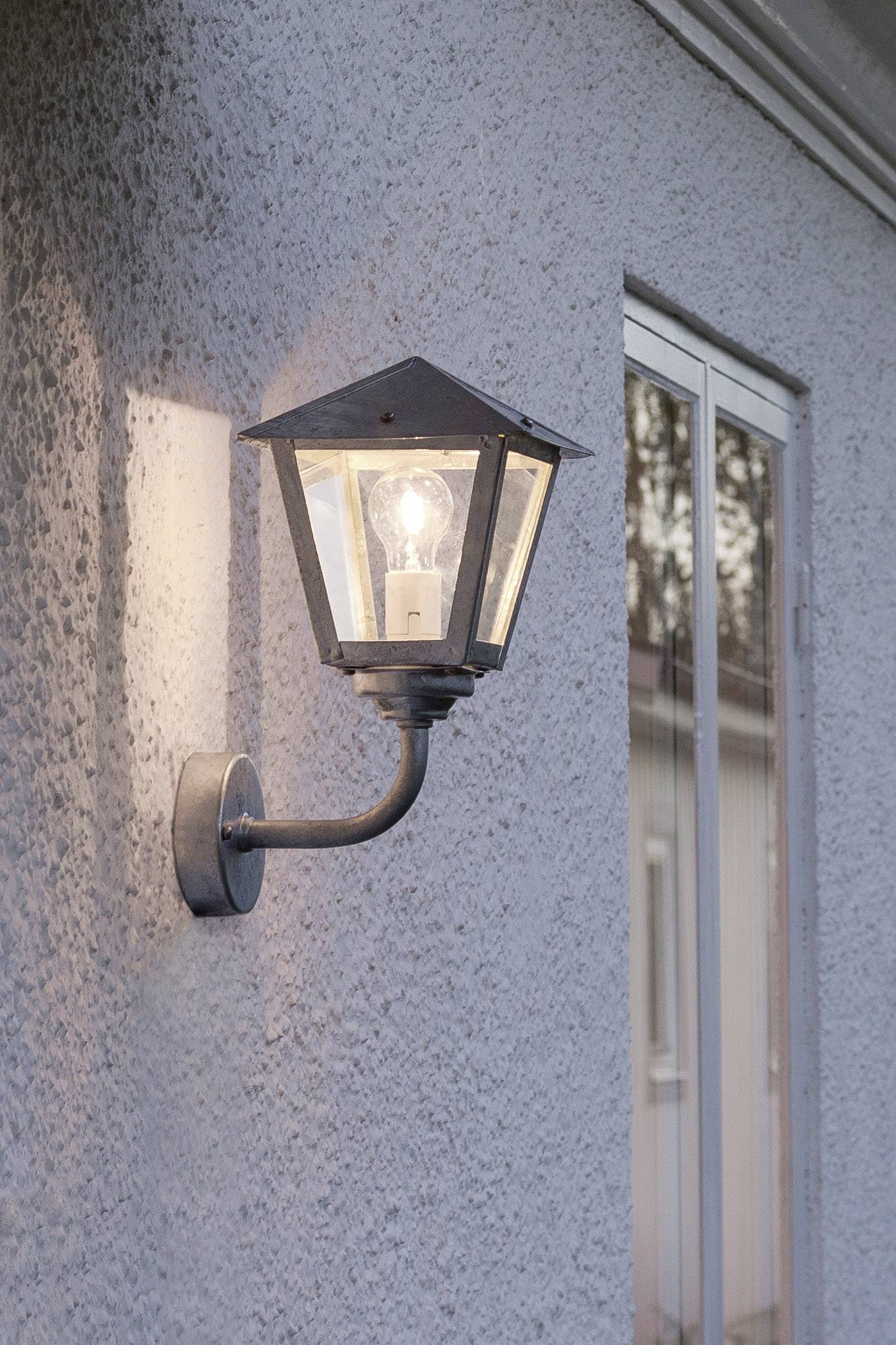 Zidna lampa na sivom zidu, upaljena u sumrak, osvjetljava prostor pored prozora.
