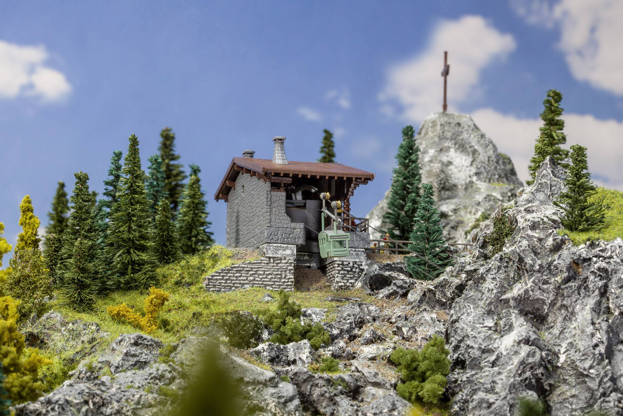 Planinska koliba u alpskom krajoliku okružena drvećem i stijenama, križić na vrhu gore nalazi se u pozadini pod vedrim plavim nebom.