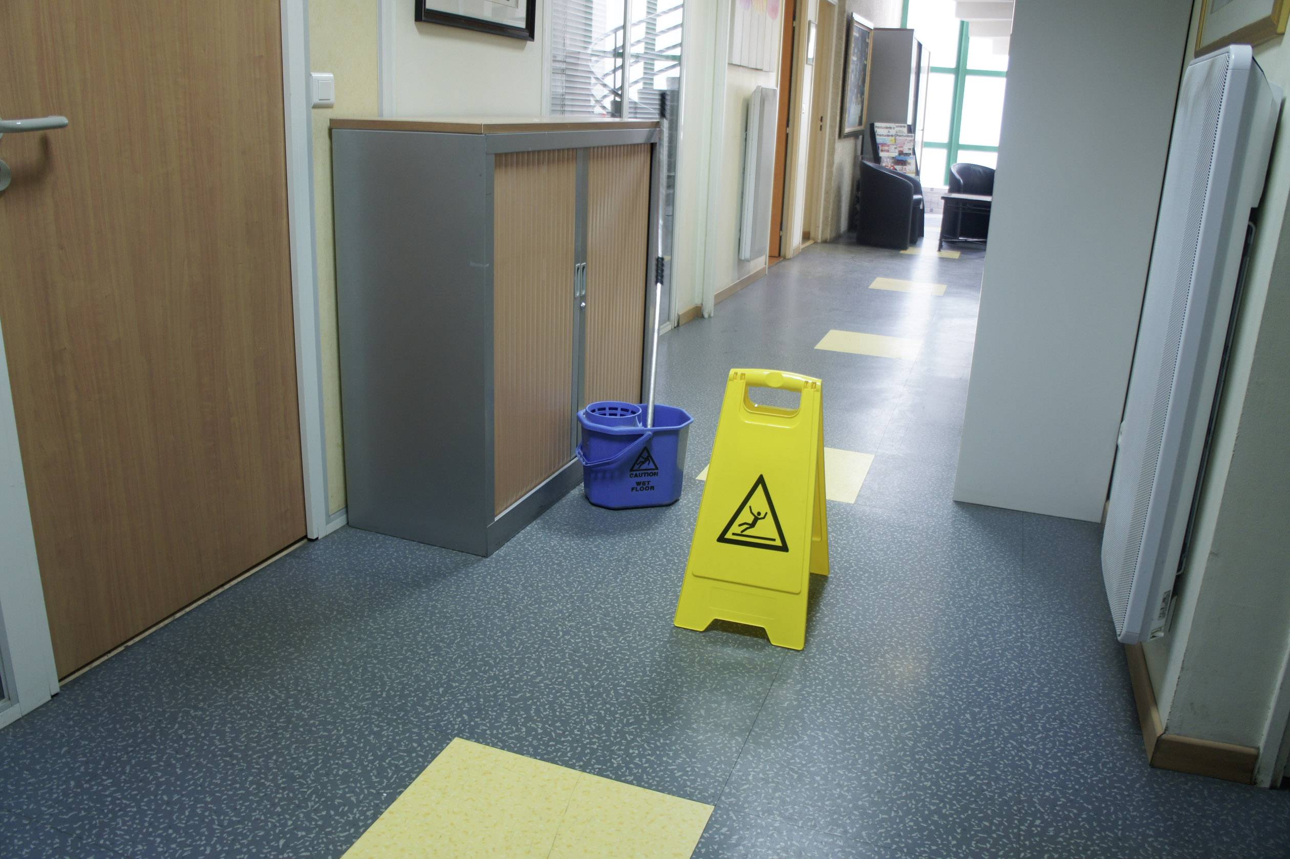 Žuti znak upozorenja s natpisom 'Opasnost od klizanja' stoji na hodniku pored plavog čistačkog koferta.