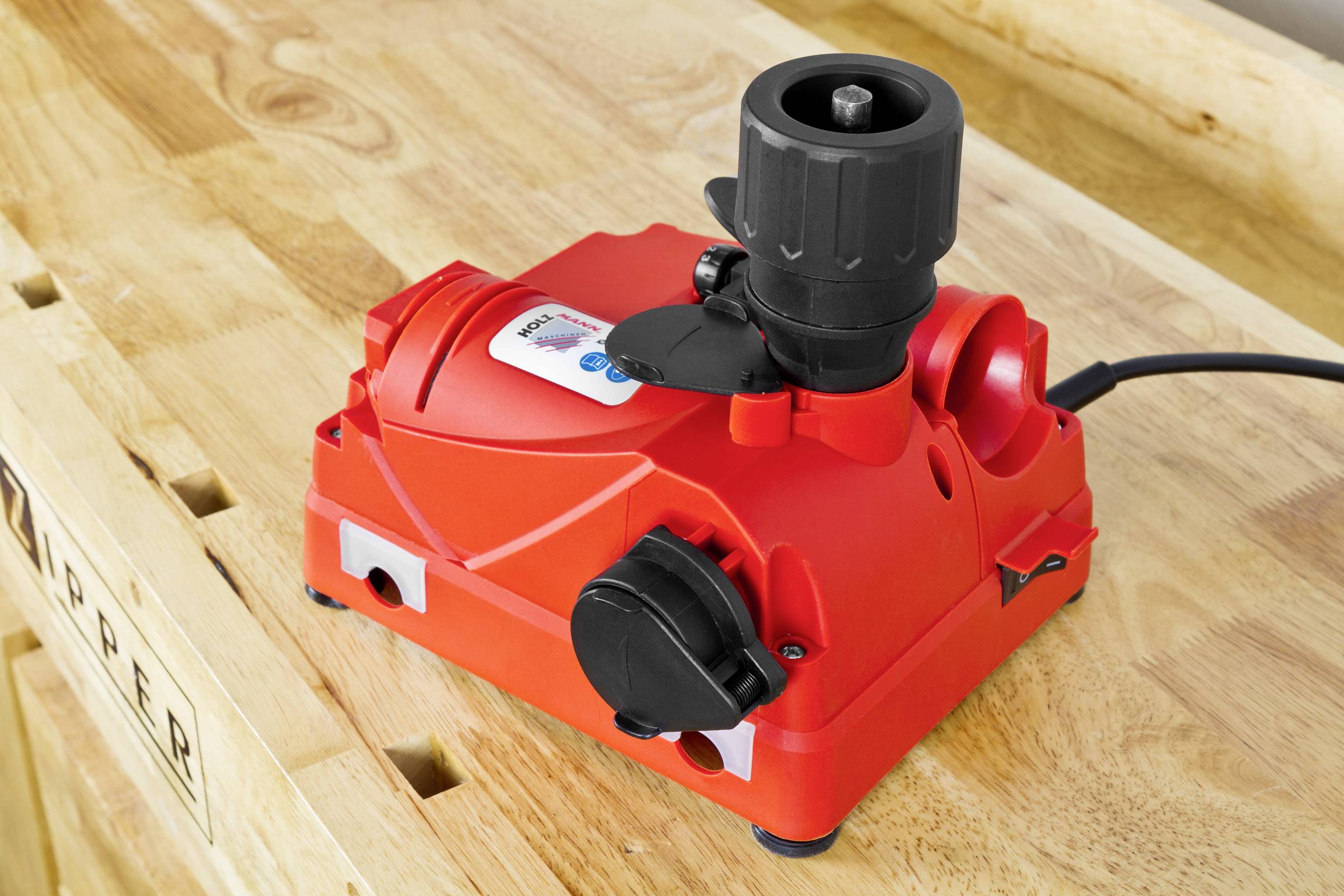 Crveni naoštrač svrdala stoji na drvenom radnom prostoru. Uređaj ima crne okretne gumbe i priključak za mrežni kabel.