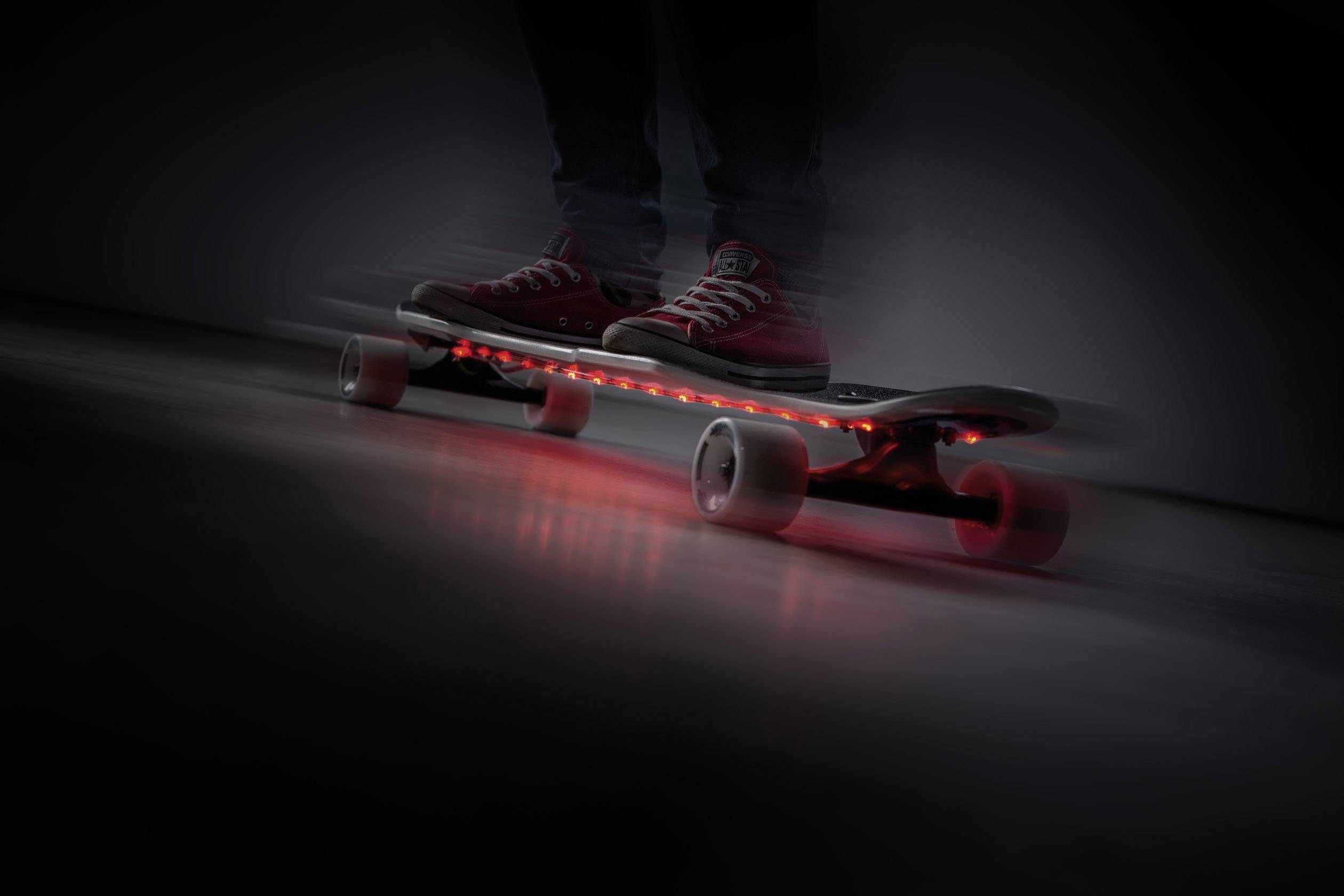 Osoba stoji na osvjetljenom skateboardu koji sa strane ima crvena svjetla, u mračnoj prostoriji.