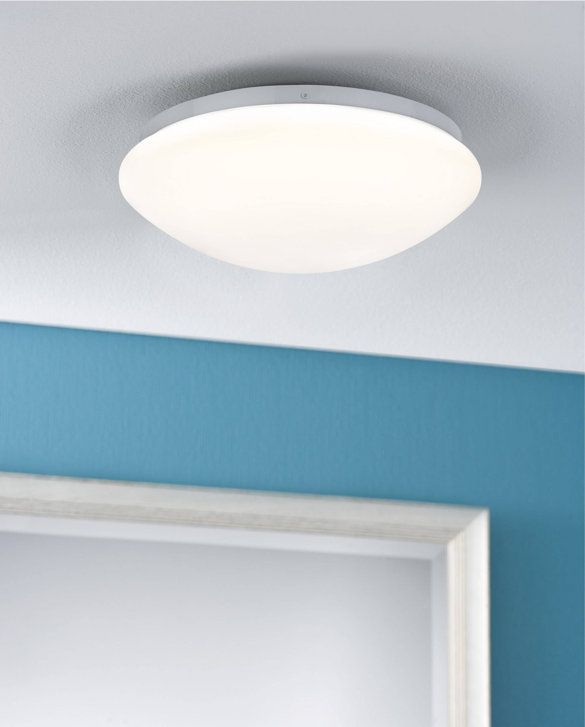 Okrugla, moderna stropna lampa emitira toplo svjetlo u prostoriji s tirkiznom bojom zida i svijetlim rubom vrata.