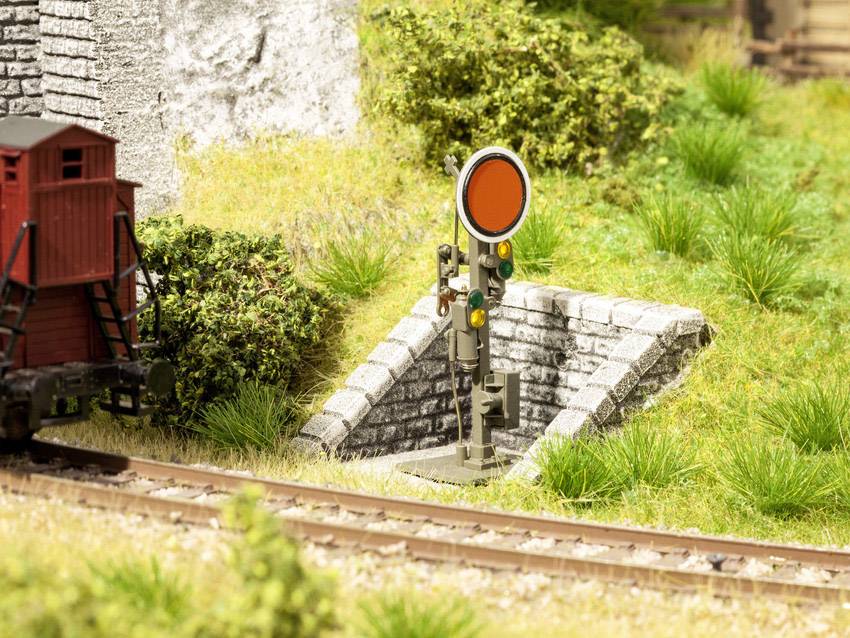 Crveni željeznički signal stoji pokraj željezničkih tračnica u miniaturnoj krajoliku, okružen zelenom vegetacijom i kućicom od opeke.