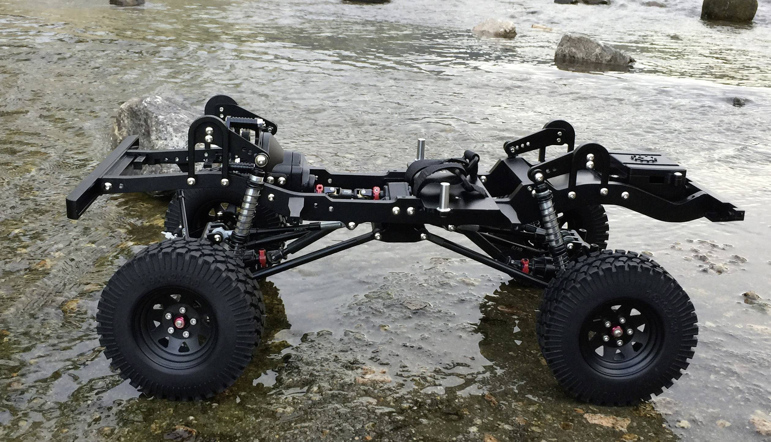 Crni model automobila s velikim gumama stoji na stijenastom tlu uz obalu tekuće vode.