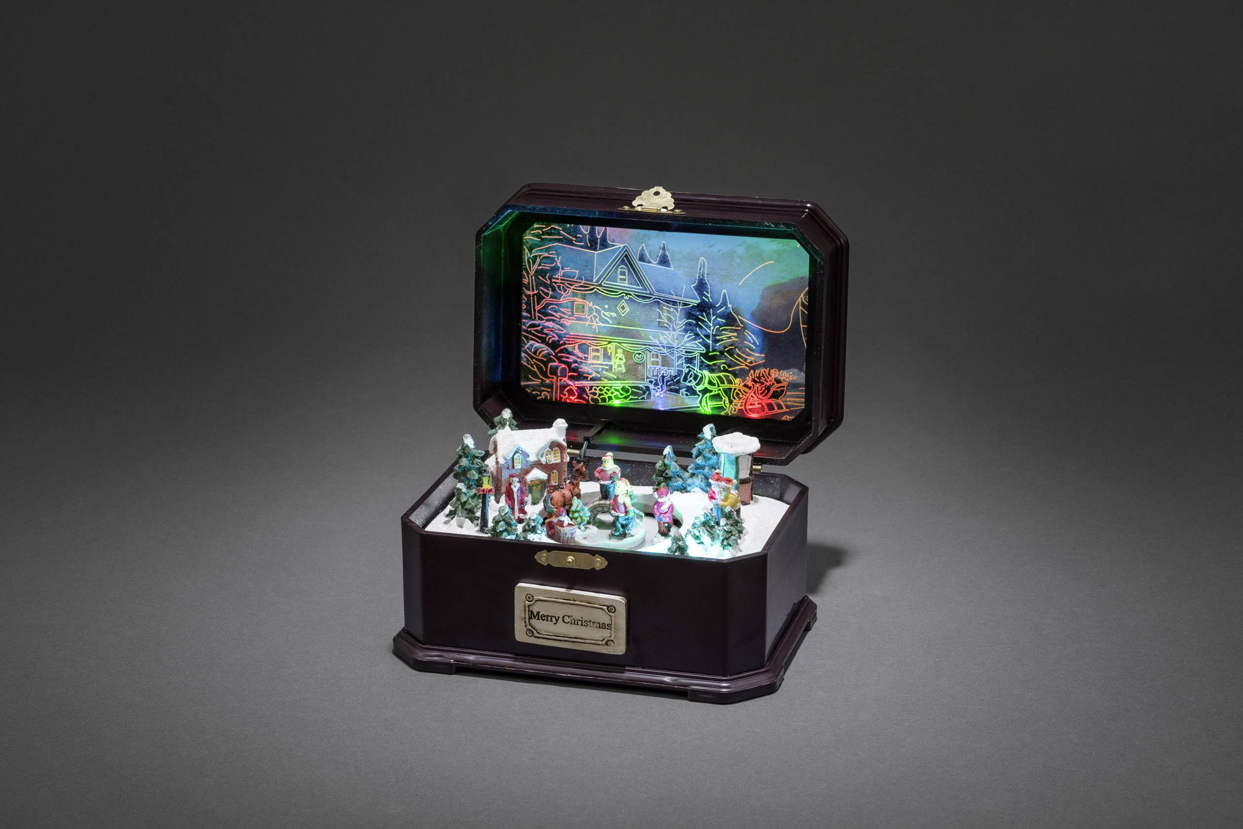 Otvorena glazbena kutija prikazuje zimsku scenu seoskog naselja s figurama i jelama, a u pozadini se svjetluca slika kuće pokrivene snijegom.