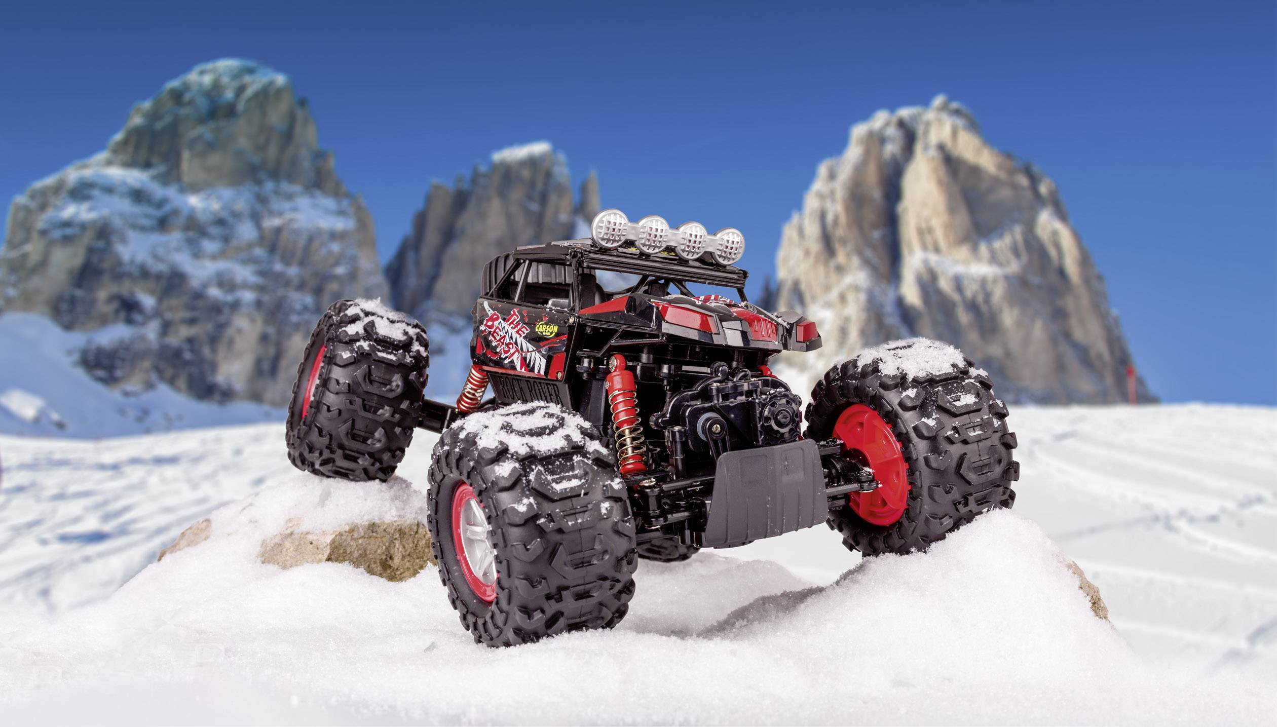 Crno-crveni modelni monster truck igračka stoji na zasnježenom terenu ispred tri istaknute stijene u pozadini.