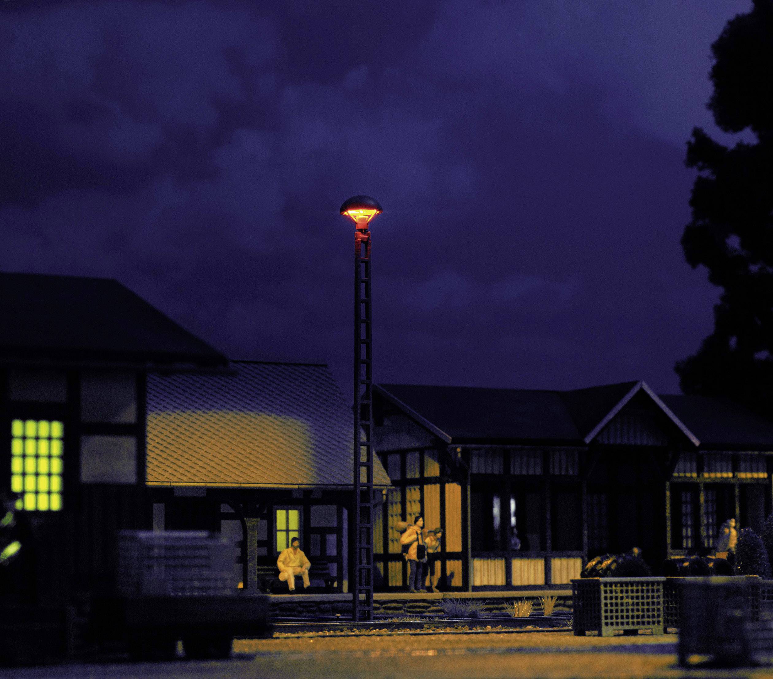 Noćna scena s osvjetljenom željezničkom stanicom u minijaturnom formatu. Ljudi čekaju ispod osvijetljene ulične lampe.