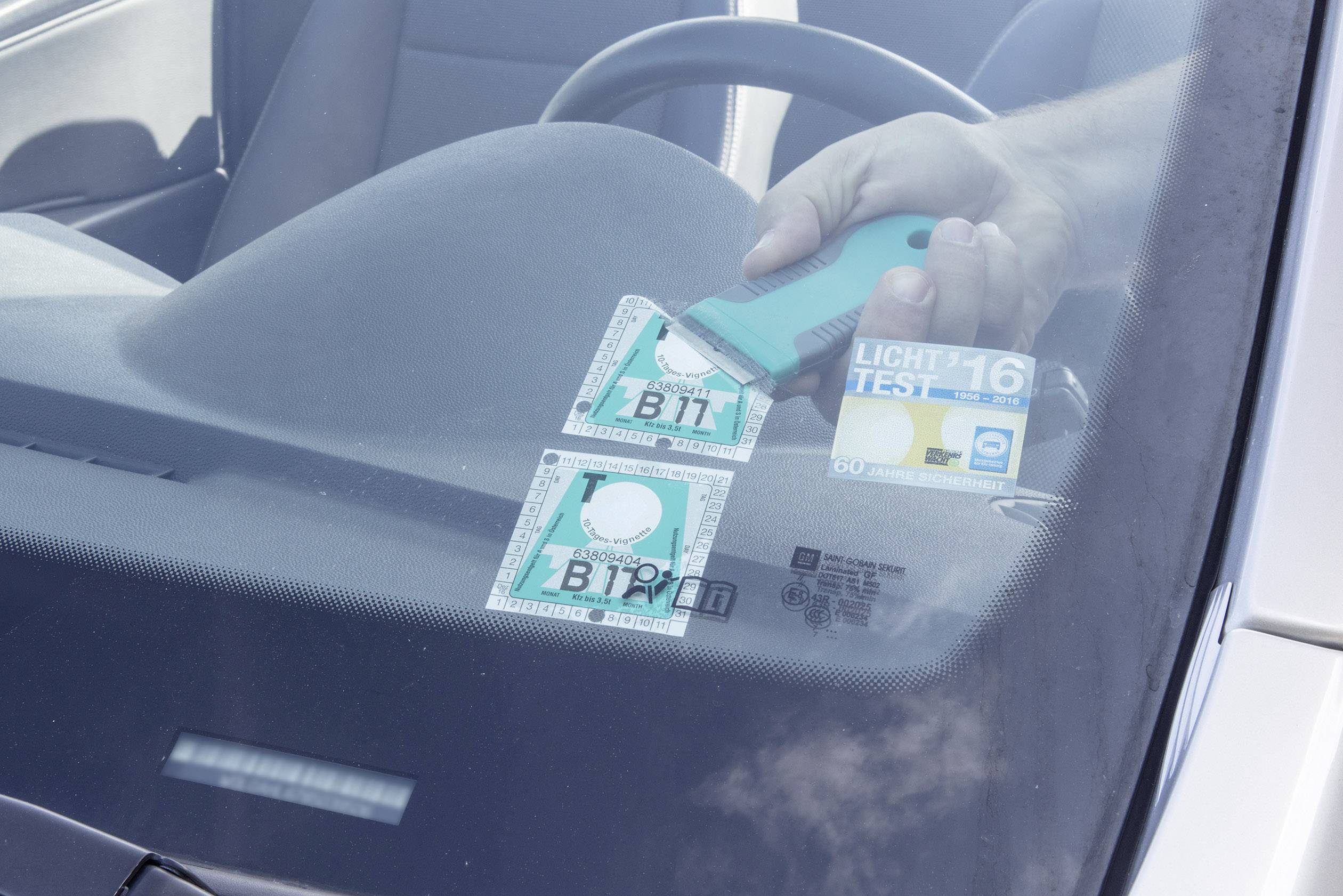 Jedna ruka pomoću alata uklanja naljepnicu za okoliš s prednjeg stakla automobila.