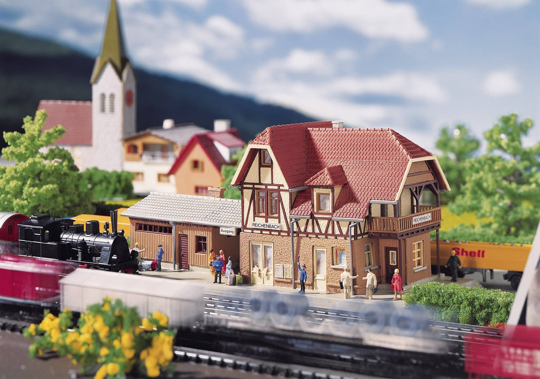 Miniaturni model njemačkog sela sa zunskom kućom, vlakom i crkvom u pozadini. Ljudi stoje ispred kuće.
