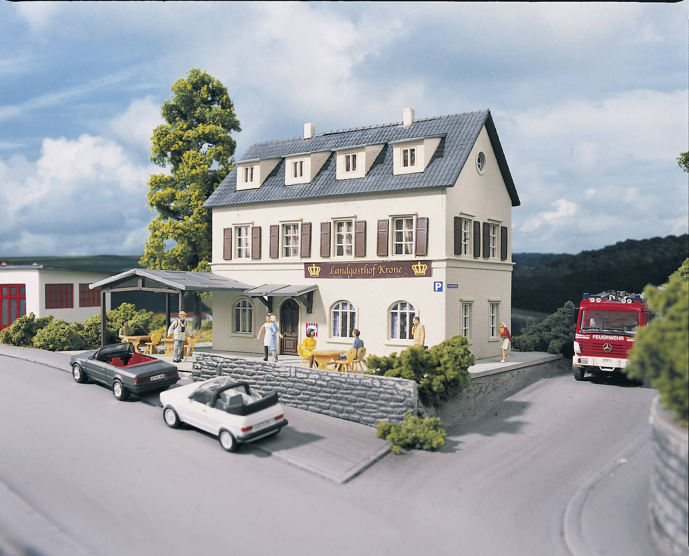 Miniaturni model dvokatne gostinice pod imenom 'Landgasthof Krone' na prometnoj raskrižnici s parkiranim automobilima i vatrogasnim vozilom.