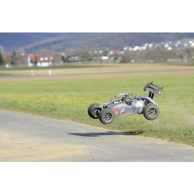 Daljinski upravljani automobil skače preko popločanog puta u ruralnom krajoliku s livadama i brdima u pozadini.