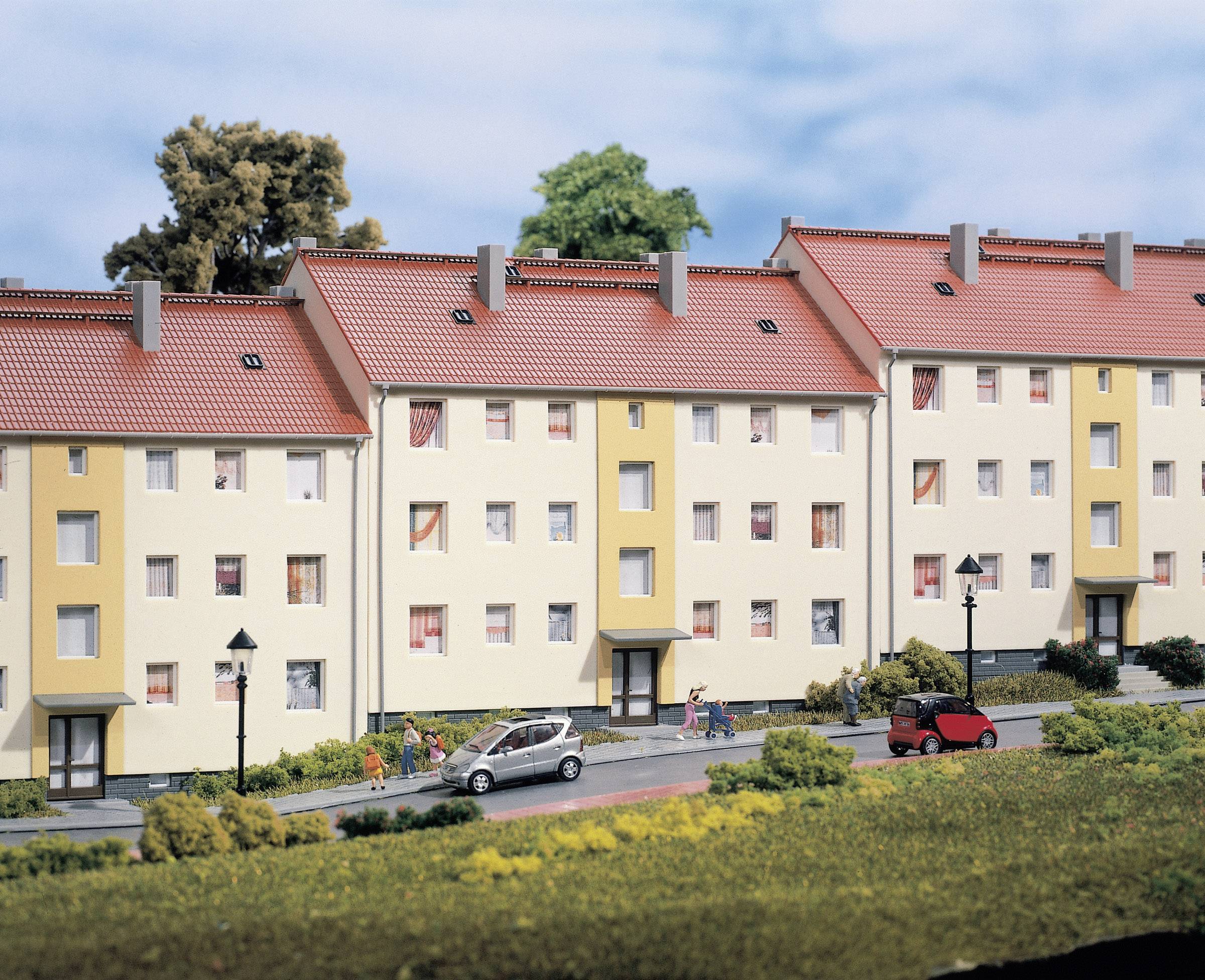 Višestambene kuće s crvenim krovovima, okružene drvećem i cestom. Automobili su parkirani, ljudi šetaju uz pješački put.