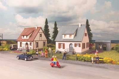 Miniaturna krajolika prikazuje dvije kuće uz cestu s malenim figurama, uključujući osobu na romobilu i automobil.