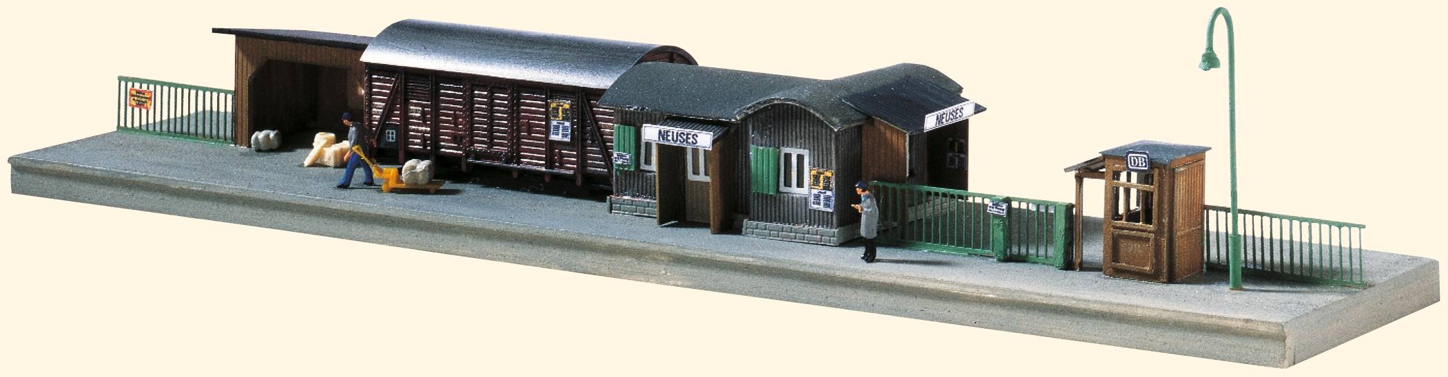 Mali model željezničke stanice s dva objekta, teretnim vagonom, figurama i ogradom na platformi.