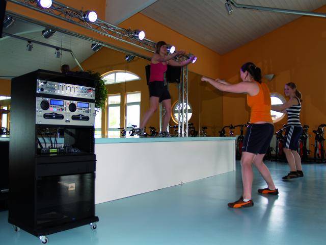 Aerobik instruktorica vodi grupu od tri osobe na fitness treningu u studiju s zrcalnim zidom i tornjem zvučnika.