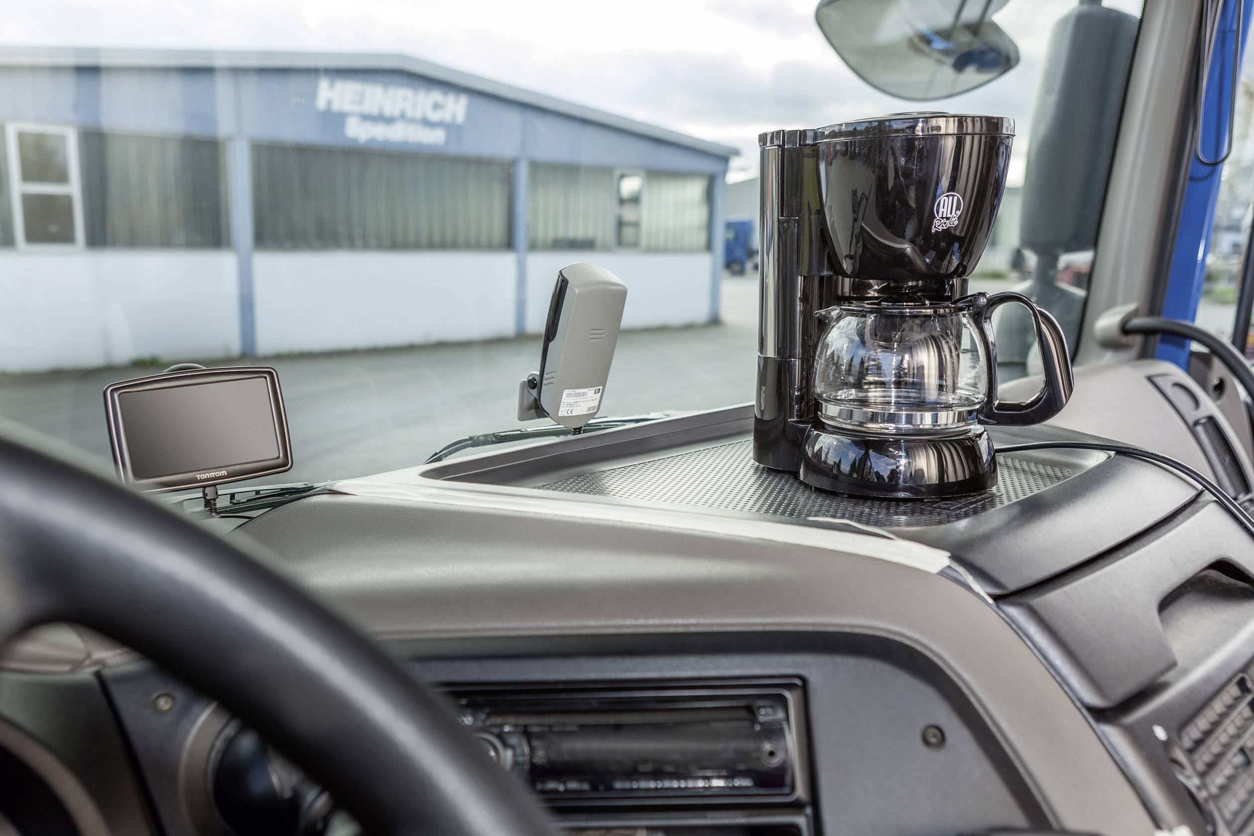 Unutar kamiona na instrumentnoj ploči stoji aparat za kavu pored navigacijskog uređaja. U pozadini se vidi skladišna hala.