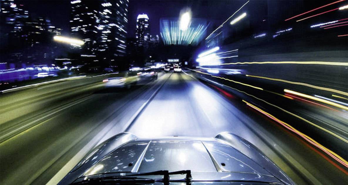 Pogled iz automobila u pokretu na noćni grad s nejasnim svjetlima koja dočaravaju brzinu i kretanje.