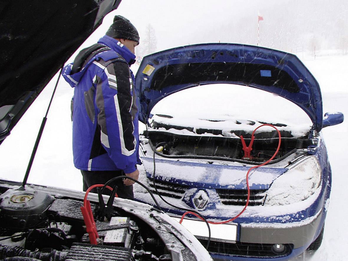 Jedna osoba pokreće automobil pomoćustarterskih kablova u snježnom okruženju. Dva automobila s otvorenim poklopcima motora su vidljiva.