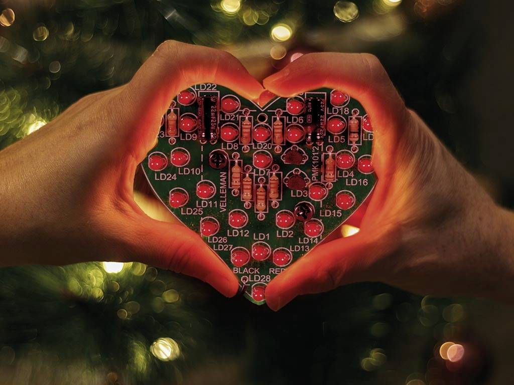 Két kéz szív alakot formál. Belül egy szív alakú, megvilágított áramkör látható piros és fekete alkatrészekkel. A háttérben fények vannak.