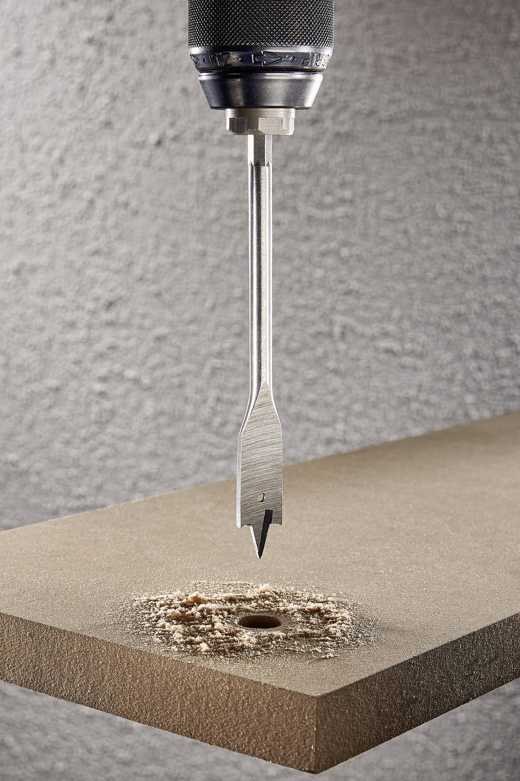 Fémfúró lyukasztja át a falemezet, forgácsokat termel. Részletfelvétel mutatja a munka precizitását szürke háttér előtt.