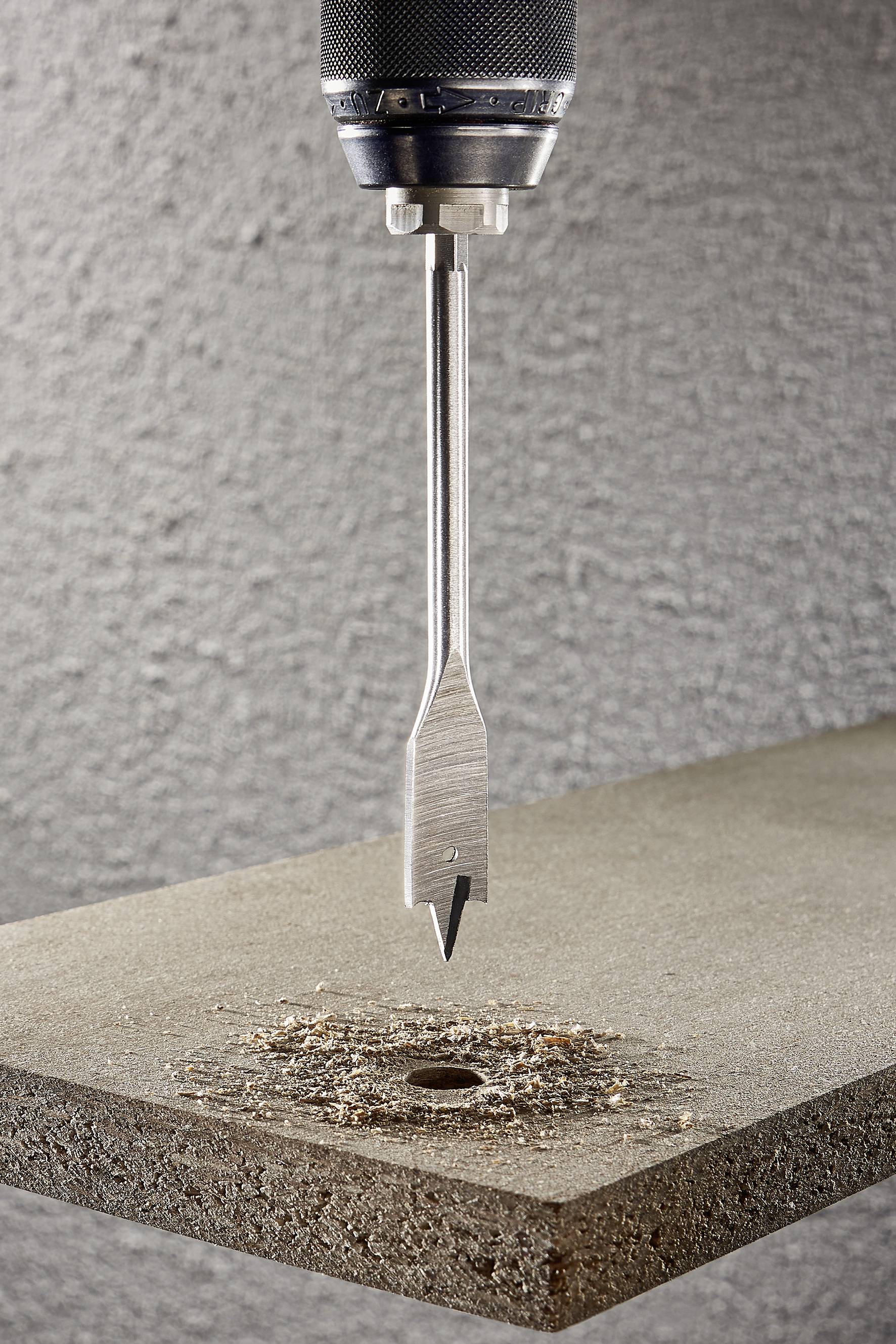 Egy fúró egy falemez felett, amelyben már van egy fúrt lyuk. Faforgácsok a lyuk körül. A háttér egy szürke, durva felület.