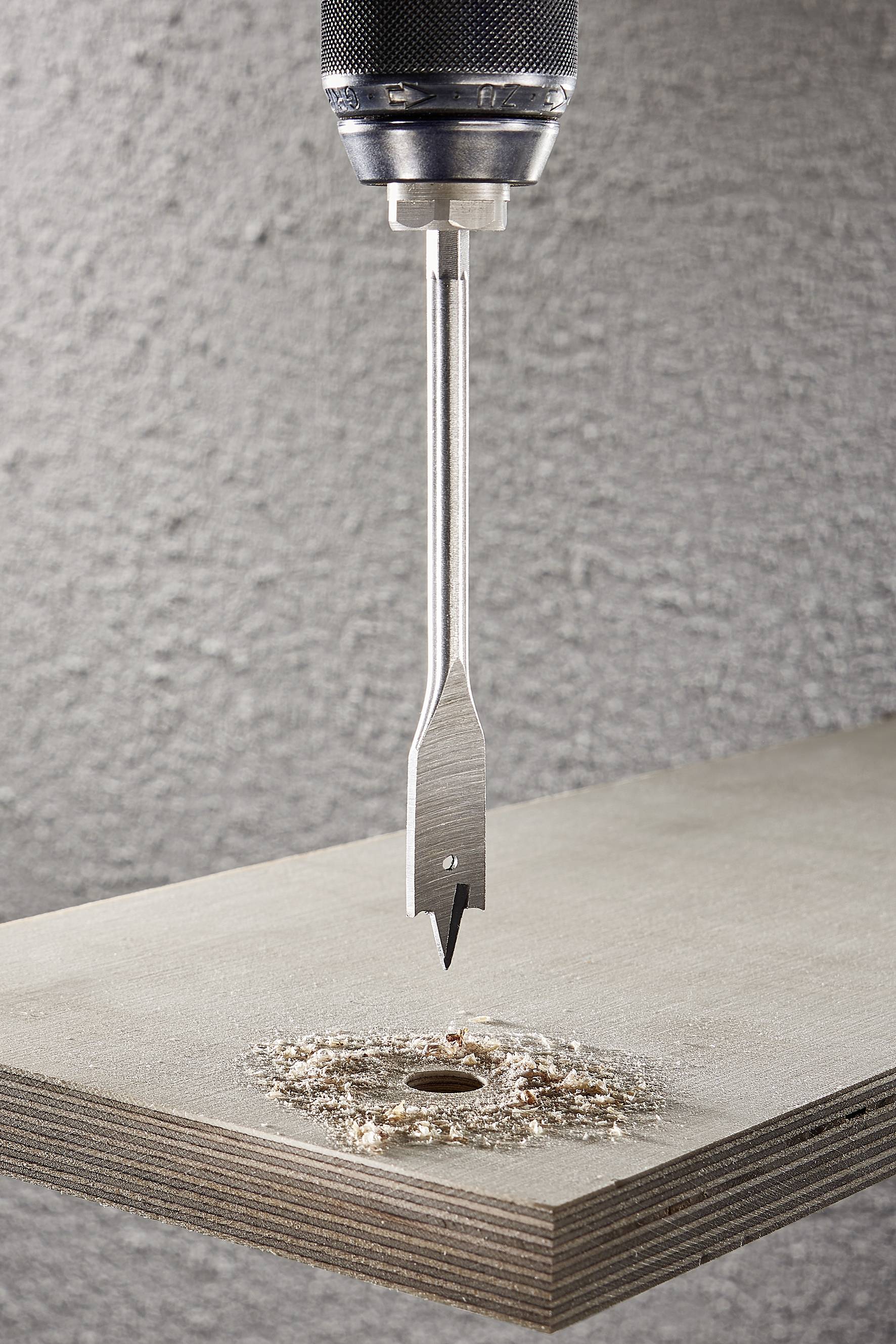 'Egy fúró közeli képe, amint lyukat fúr egy falemezbe. Faforgácsok és por szóródnak a lyuk körül.'