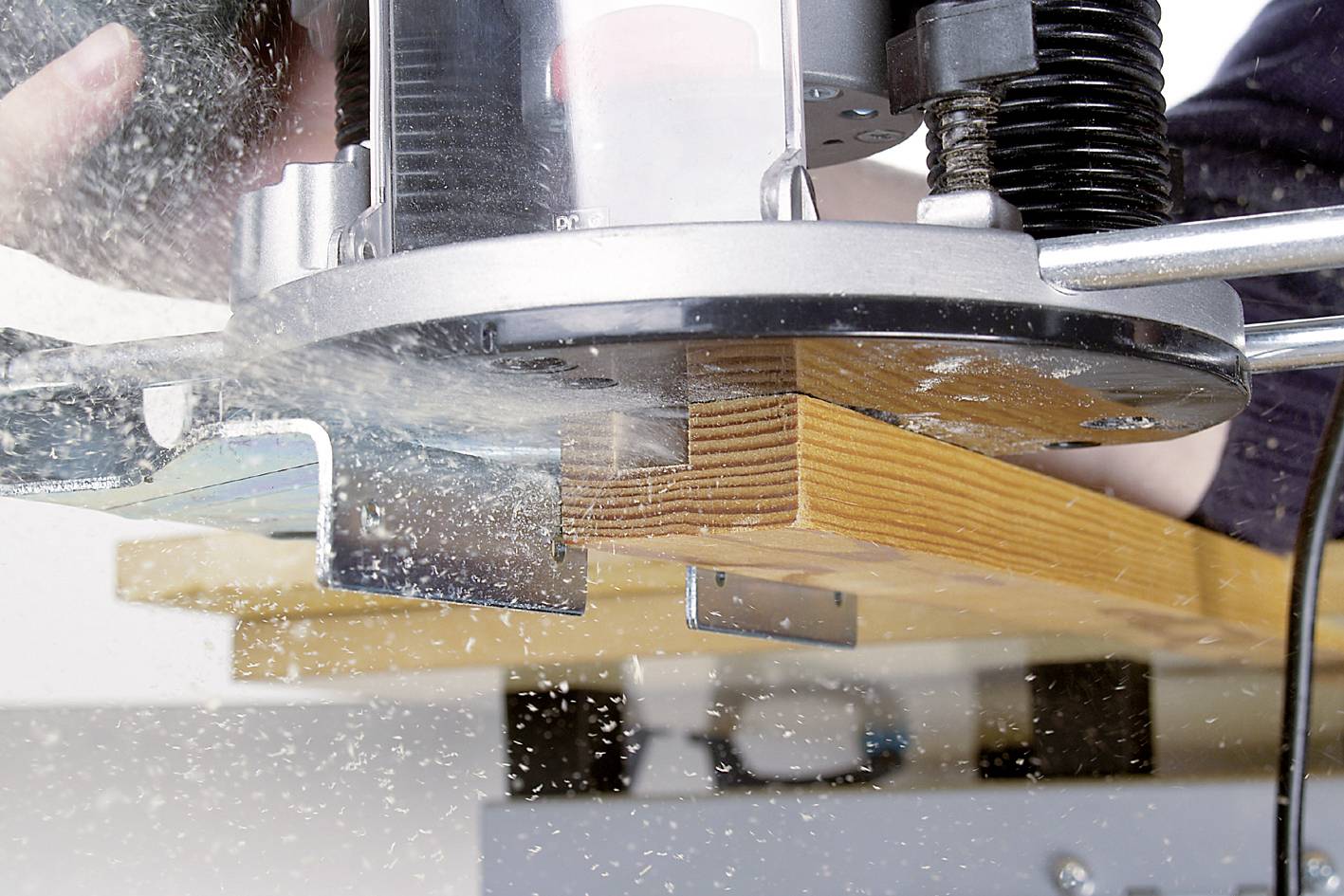 Egy felső marógép egy darab fát munkál meg. Faforgácsok repülnek a levegőben. A készüléket egy személy kezeli, a fókusz a gépen van.