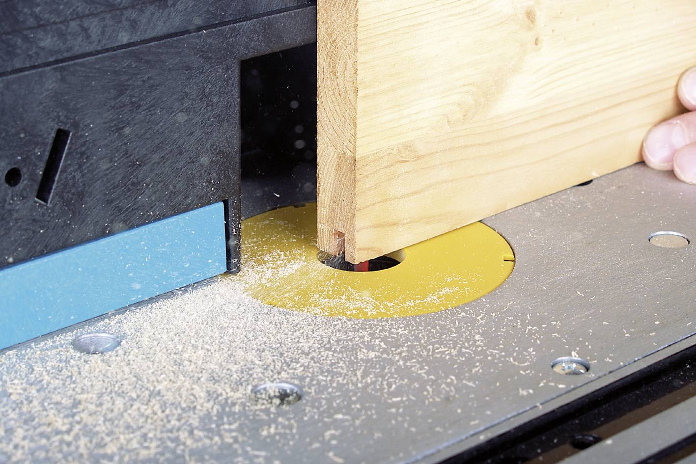 Egy falemez megmunkálása történik egy asztali marógépen. Faforgácsok vannak szétszórva a szerszám körül. Egy kéz tartja a lemezt.