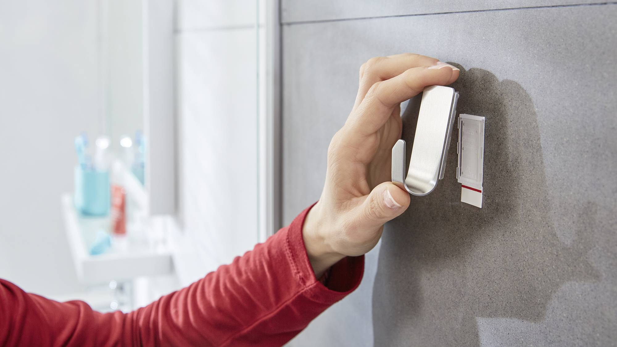 Egy piros ujjú kéz egy kampót vesz le egy szürke falról. A háttérben fürdőszobai eszközök láthatók egy polcon.