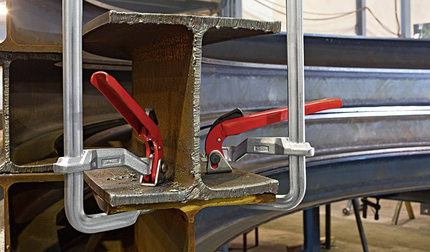 Két fémtartót nagy, piros fogókarú szorítók tartanak össze. A környezet egy műhelyre emlékeztet.