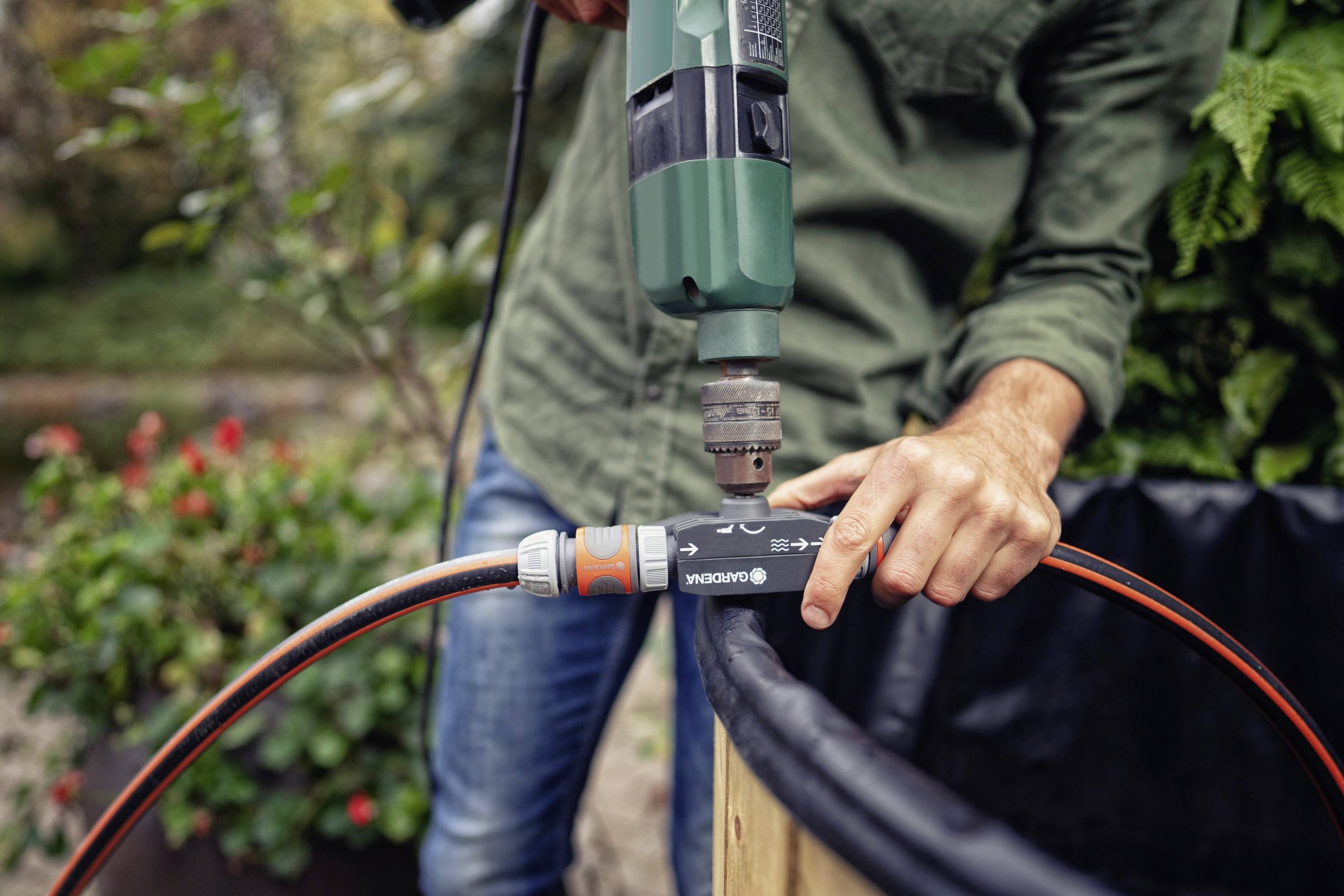 Egy zöld inget viselő személy fúrógépet használ, hogy rögzítsen egy csatlakozót egy kerti tömlőn. A háttérben növények láthatók.