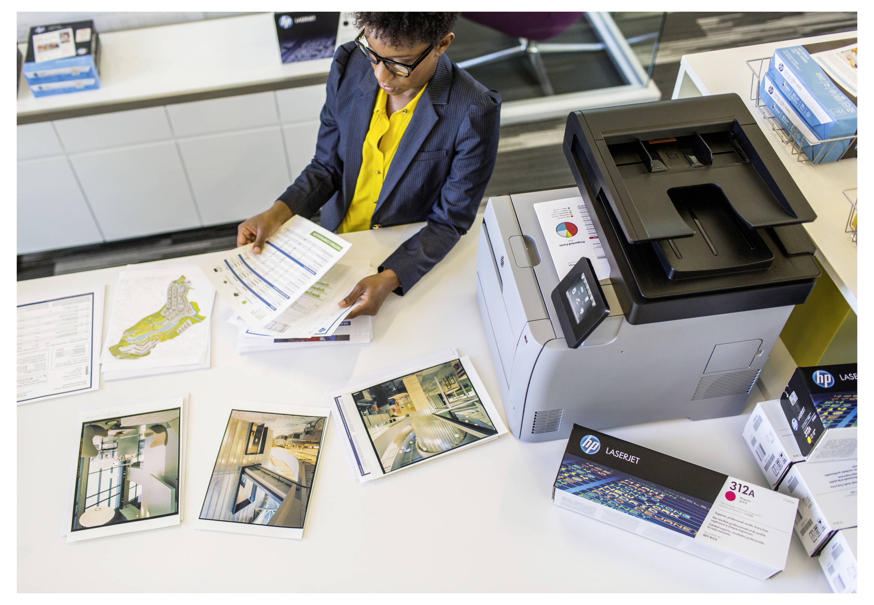 Irodai dolgozó nyomtatott dokumentumokat áttekint egy nagy HP LaserJet nyomtató mellett az asztalon, amelyen mintafotó nyomatok és szállítási dobozok találhatók.