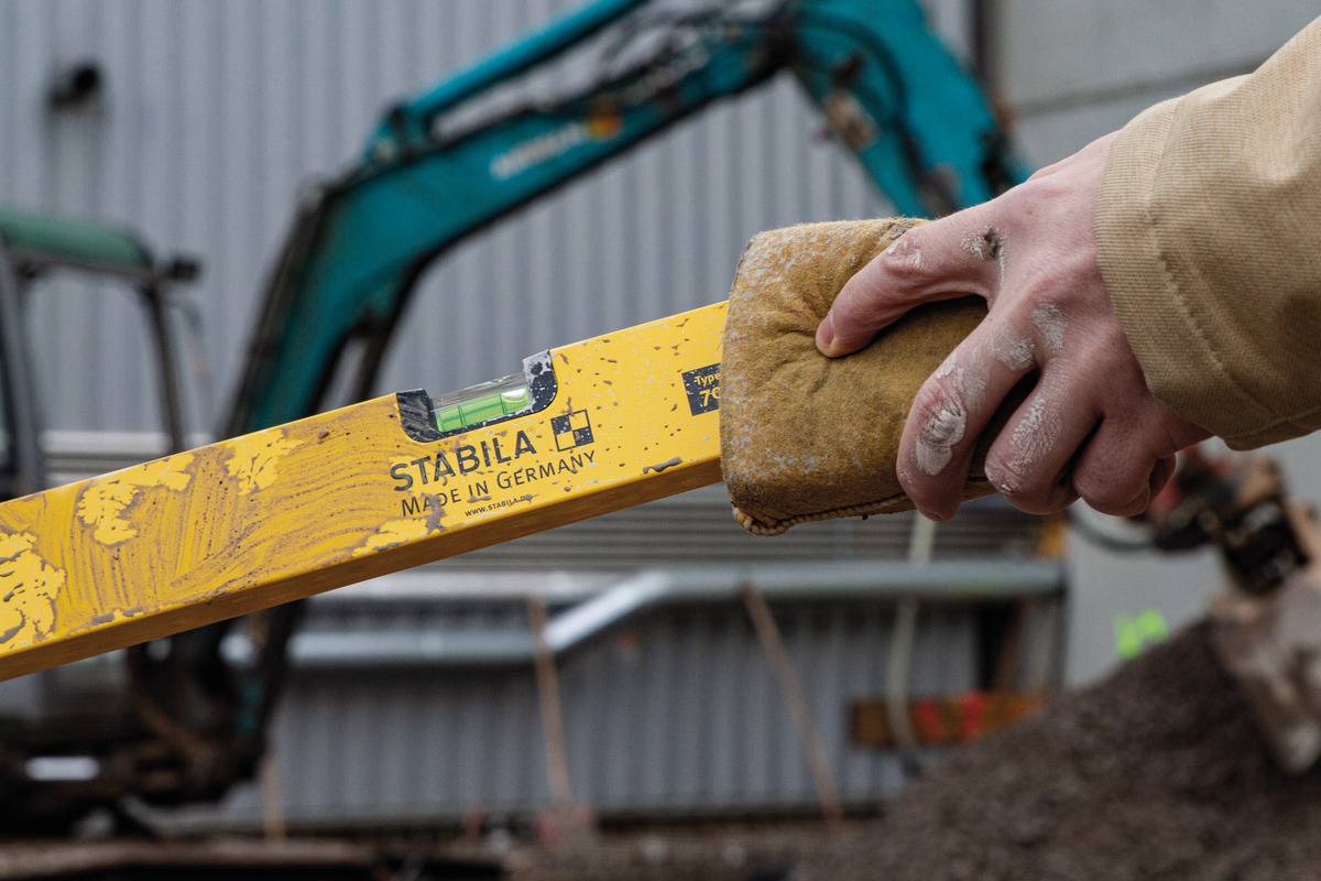Egy személy vízszintmérőt tart egy építkezésen. A háttérben egy elmosódott kotrógép látható.