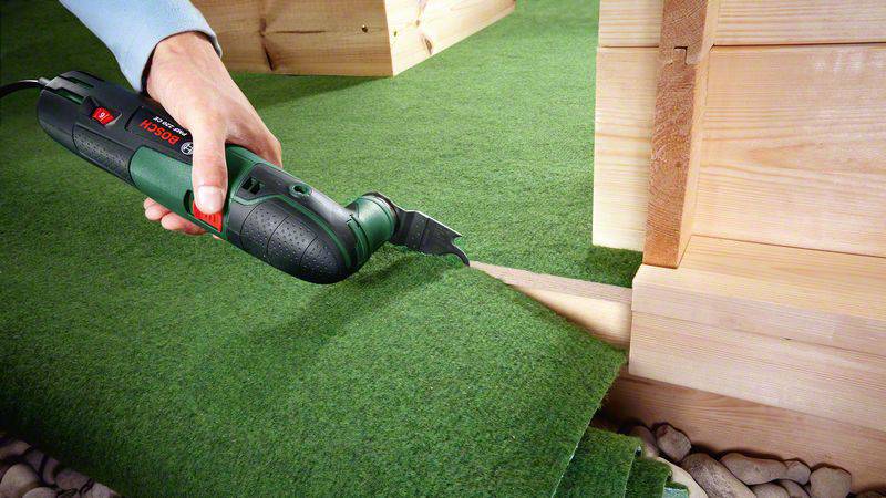 Egy személy egy elektromos szerszámmal műfüvet vág körbe egy fa épület sarkánál, hogy pontosan illeszkedjen.