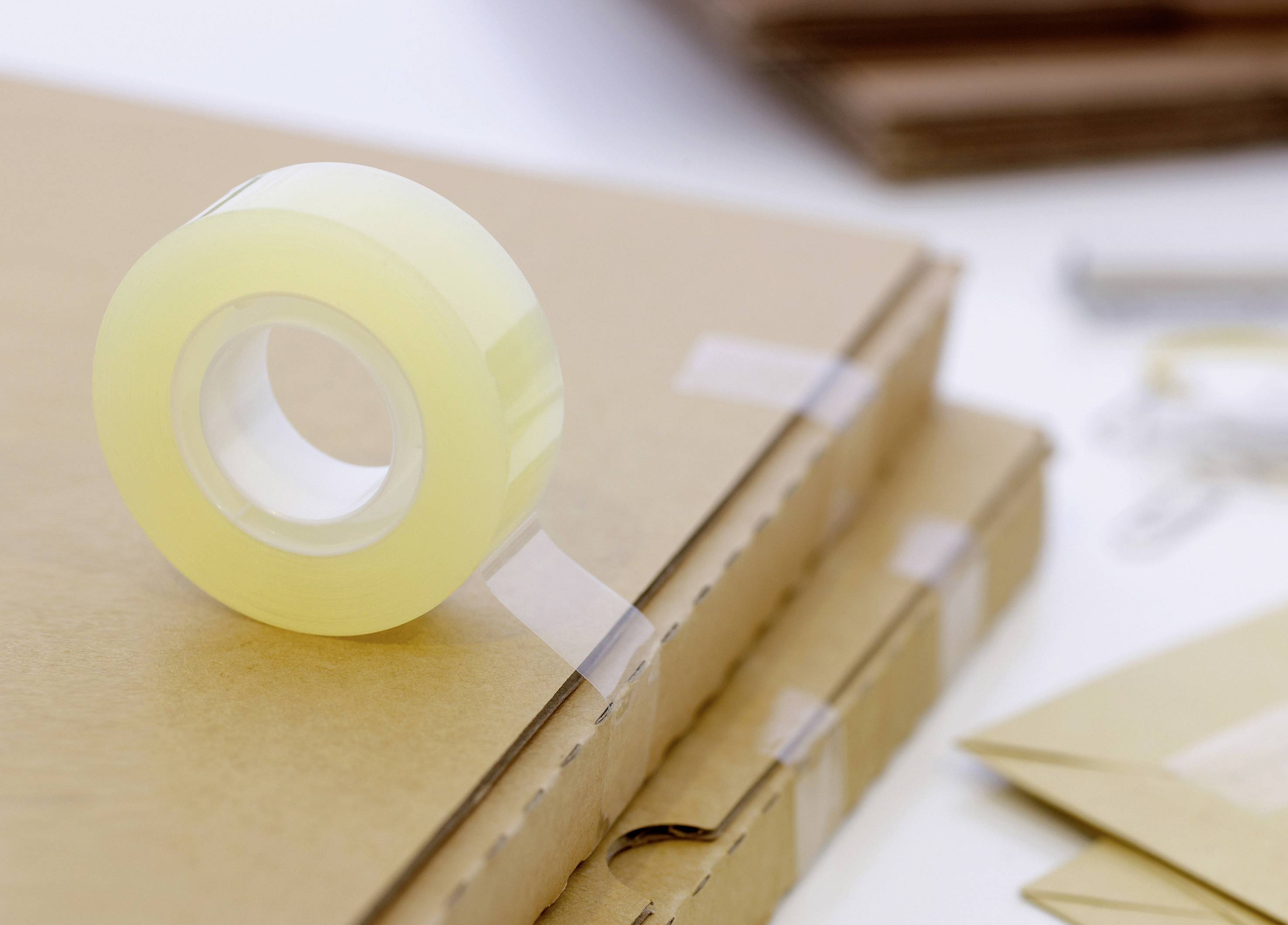 Egy tekercs ragasztószalag több egymásra pakolt doboz tetején fekszik. A háttérben gemkapcsok és borítékok láthatók.