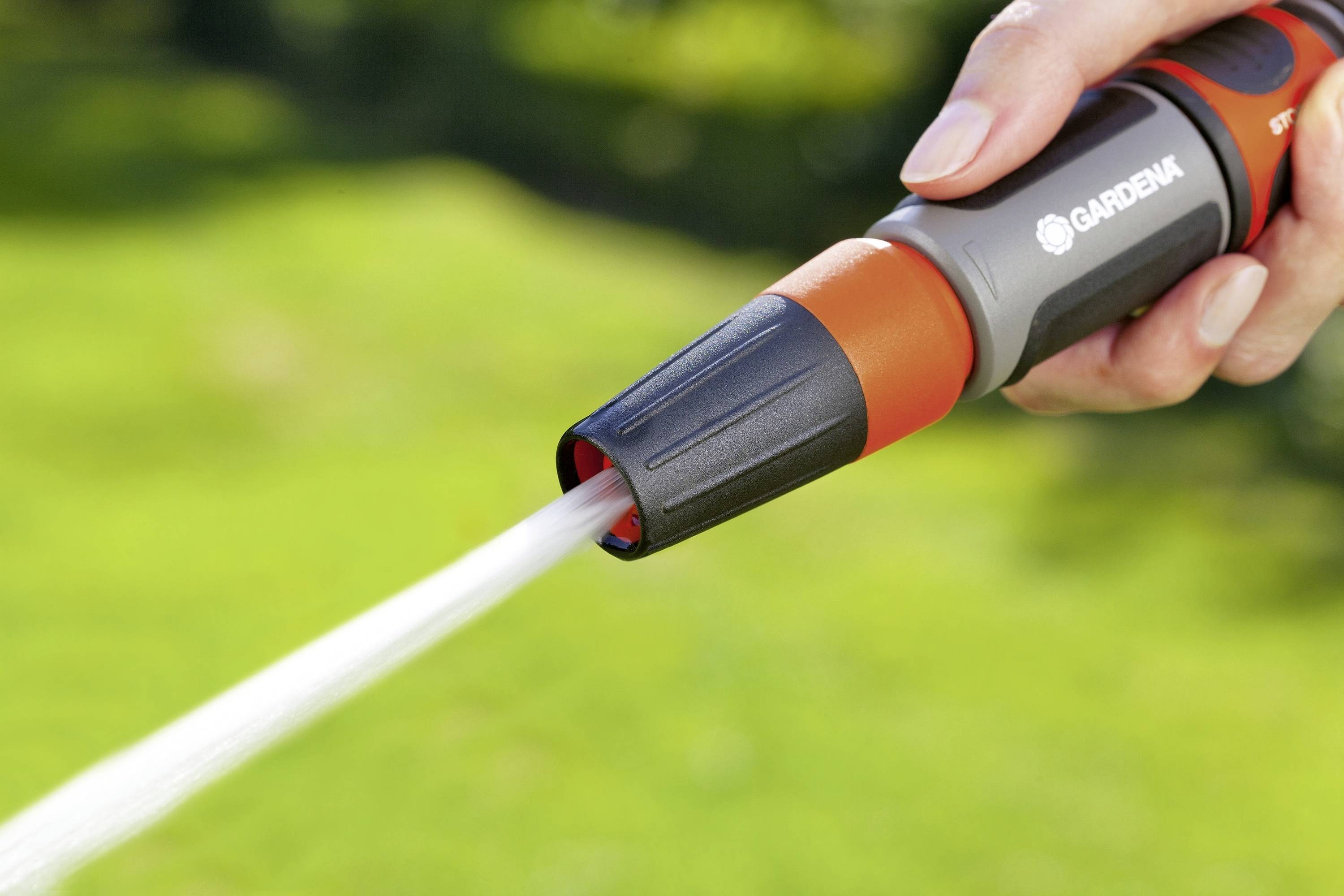 Közeli felvétel egy kézről, amely egy szórófejjel ellátott kerti tömlőt tart. A vízsugar egy zöld gyepre irányul.