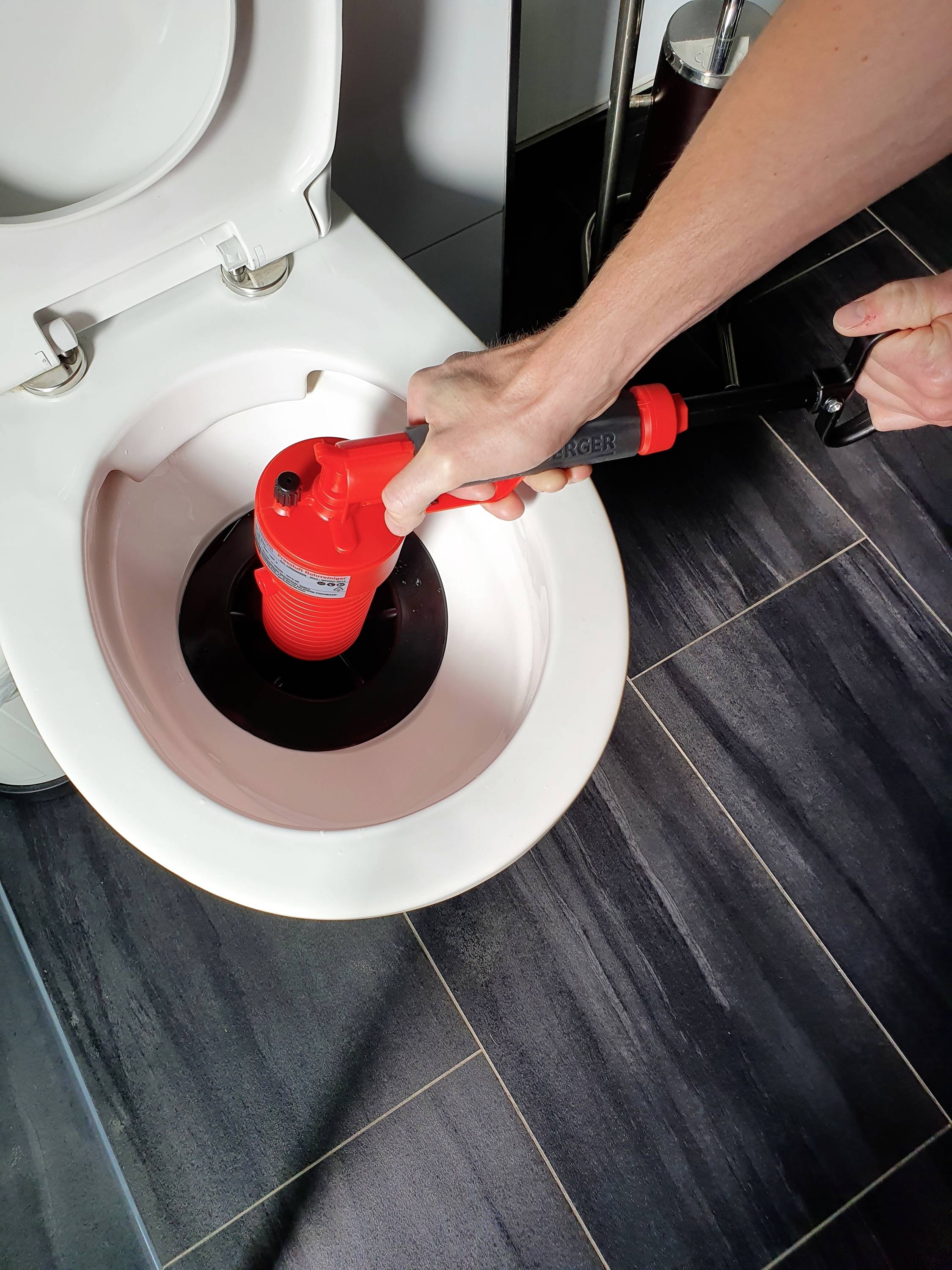 Egy kéz piros szívókorongot használ egy eldugult WC kitisztítására. A jelenet egy sötét csempével burkolt fürdőszobában játszódik.