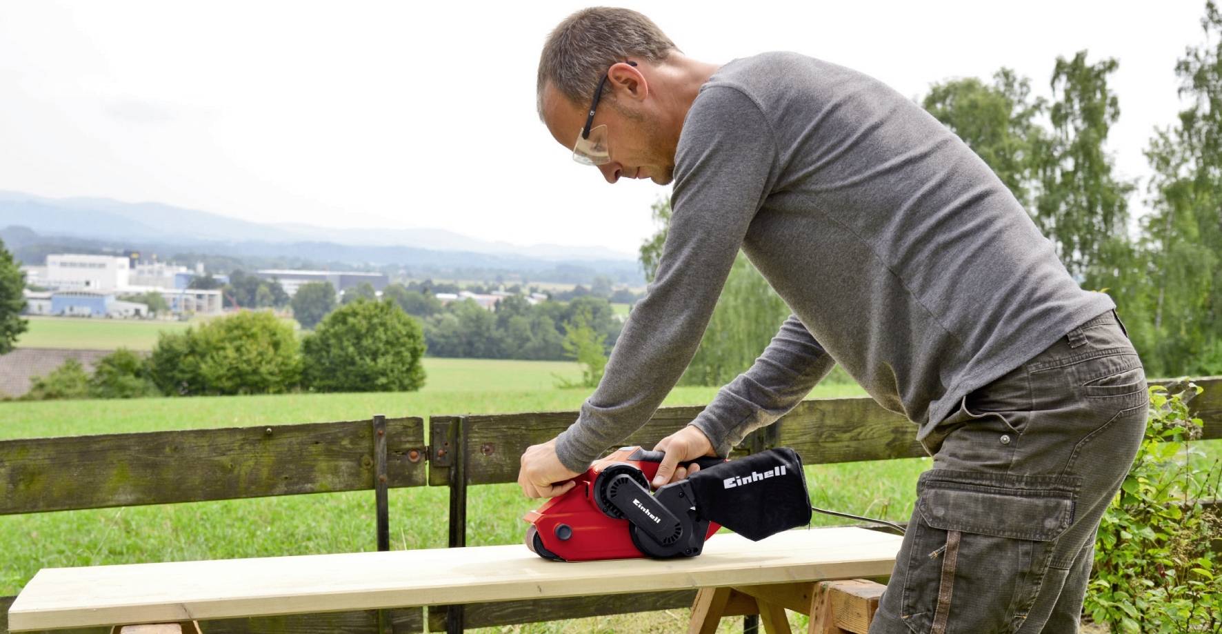 Egy férfi egy szalagcsiszológéppel csiszol egy fadeszkát a szabadban. A háttérben fák és egy városi táj látható.